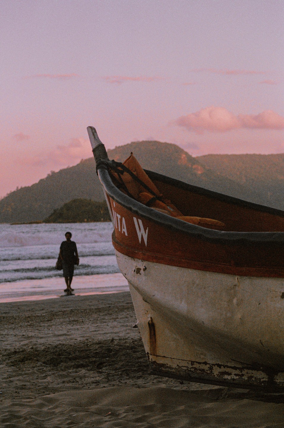 A small boat rests on a sandy beach with a person walking in the distance as the sun sets over mountains.