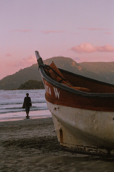 A small boat rests on a sandy beach with a person walking in the distance as the sun sets over mountains.
