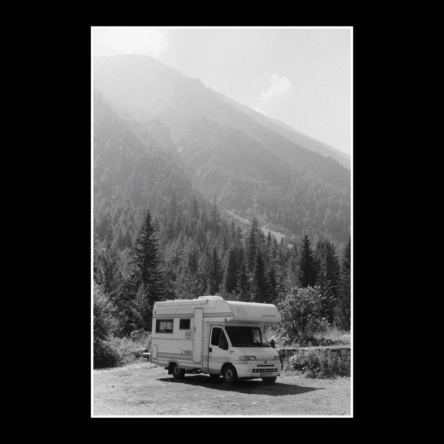 A camper van is parked on a dirt road with a forest and mountain in the background.