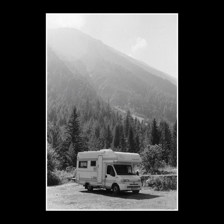 A camper van is parked on a dirt road with a forest and mountain in the background.