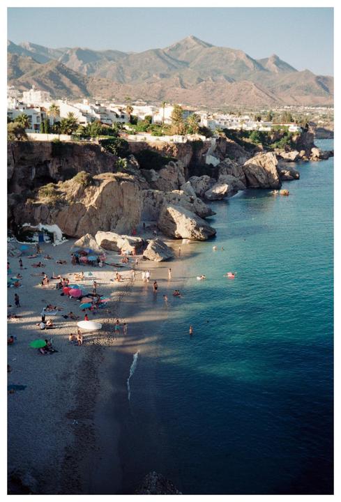 A beach with many people and colorful umbrellas is nestled below cliffs with white buildings, with mountains in the background.