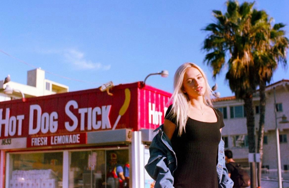 A woman stands in front of a Hot Dog on a Stick stand with palm trees and buildings in the background.