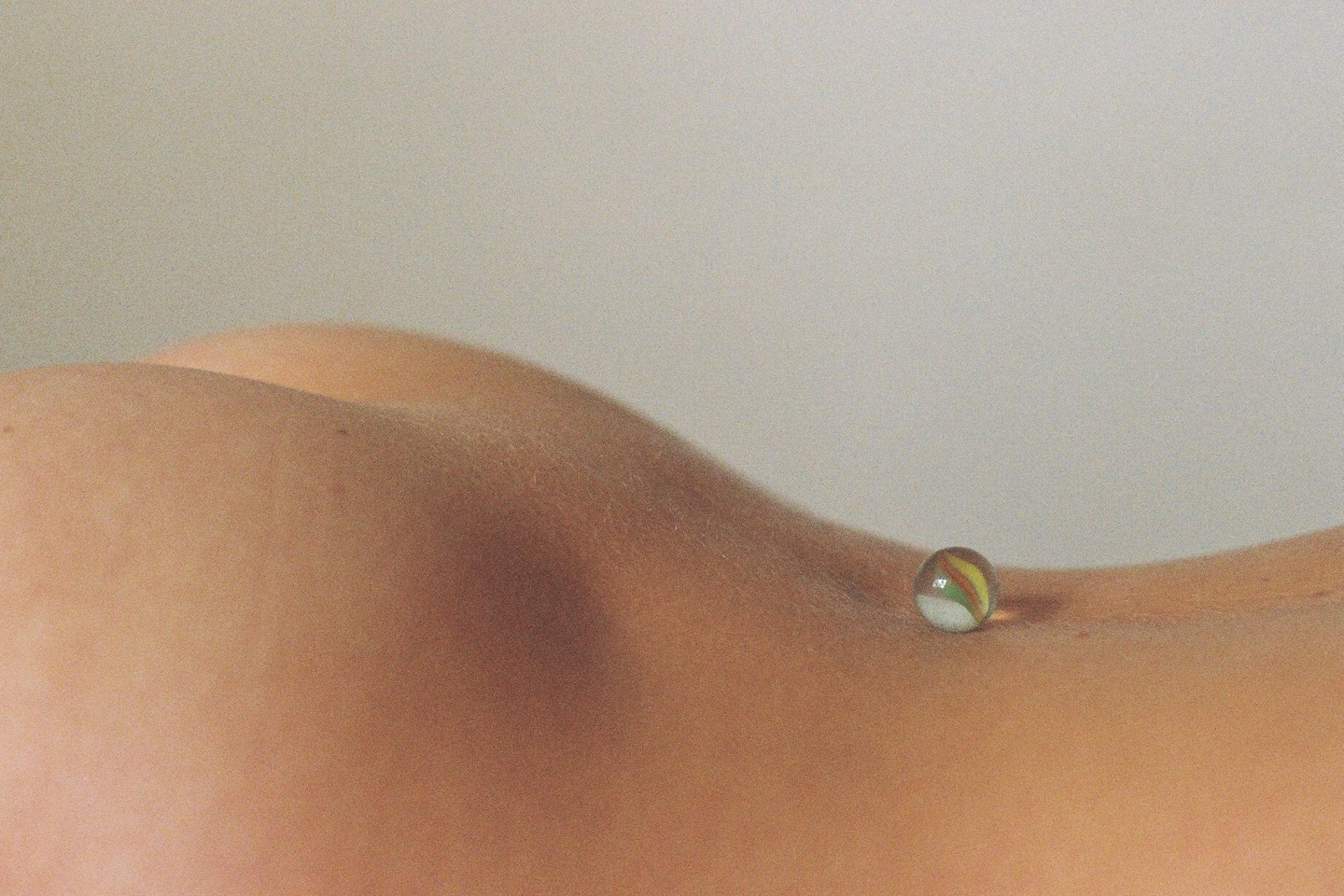 A glass marble rests on a person's skin, which forms soft curves against a plain background.