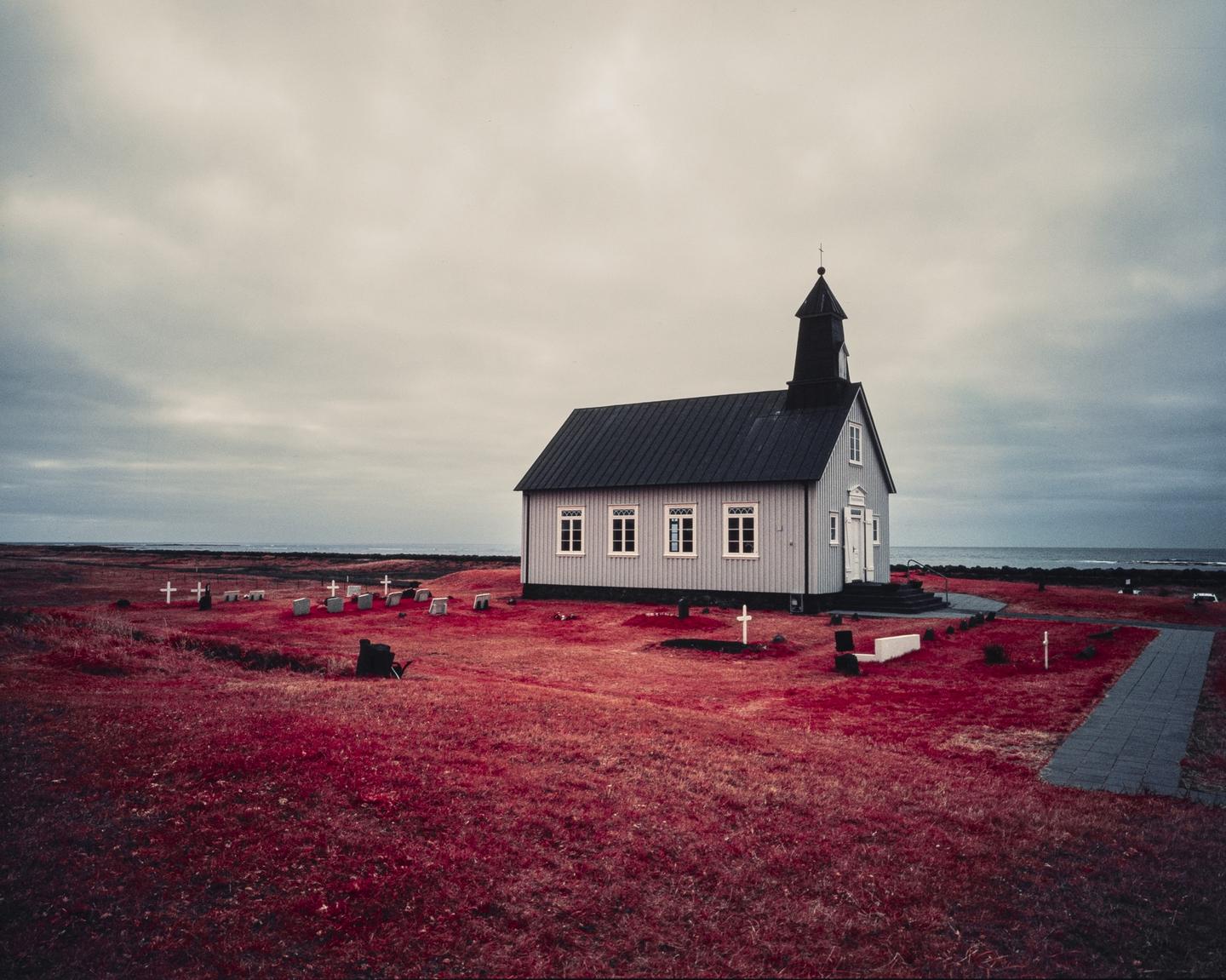 A small church with a graveyard is situated on a red grassy field next to the ocean under a cloudy sky.