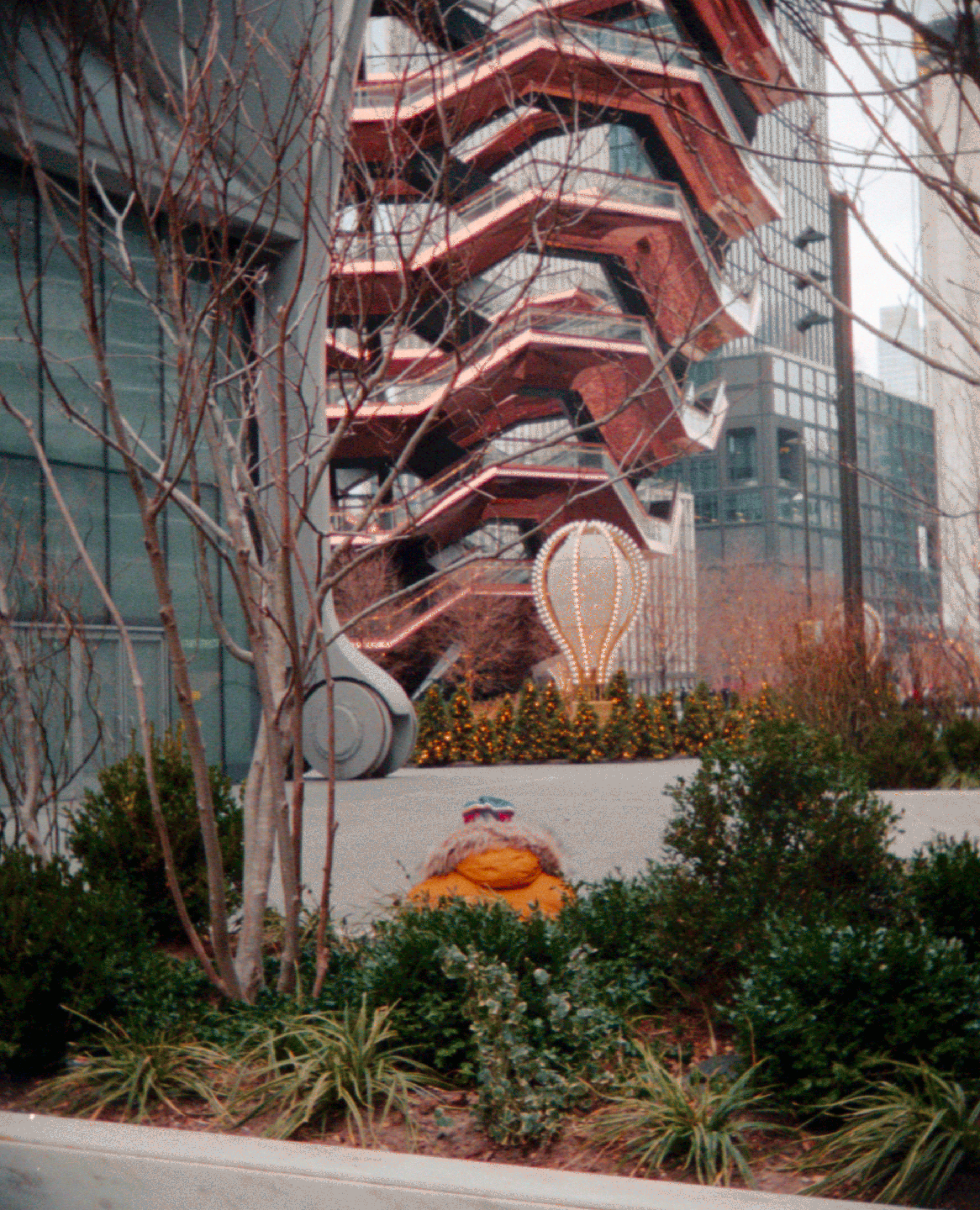 A person in a yellow coat sits among bushes with the Vessel building and a hot air balloon sculpture in the background.