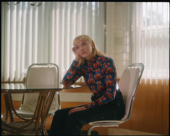 A woman with blonde hair sits at a table, resting her head on her hand and looking up.