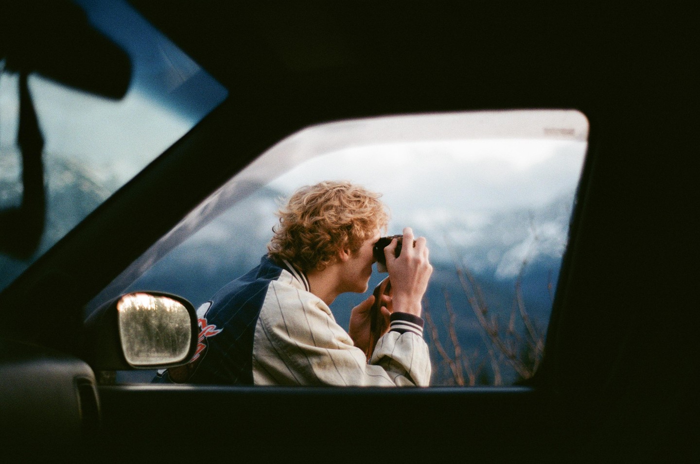 A young man with curly hair looks through a camera out of a car window at a mountain.