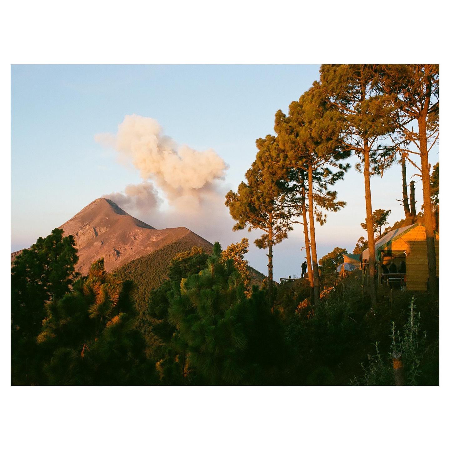A volcano emits a plume of smoke into the sky above a forest with tall trees and a small cabin.