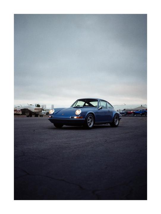 A blue car is parked on an asphalt surface with airplanes in the background under a cloudy sky.