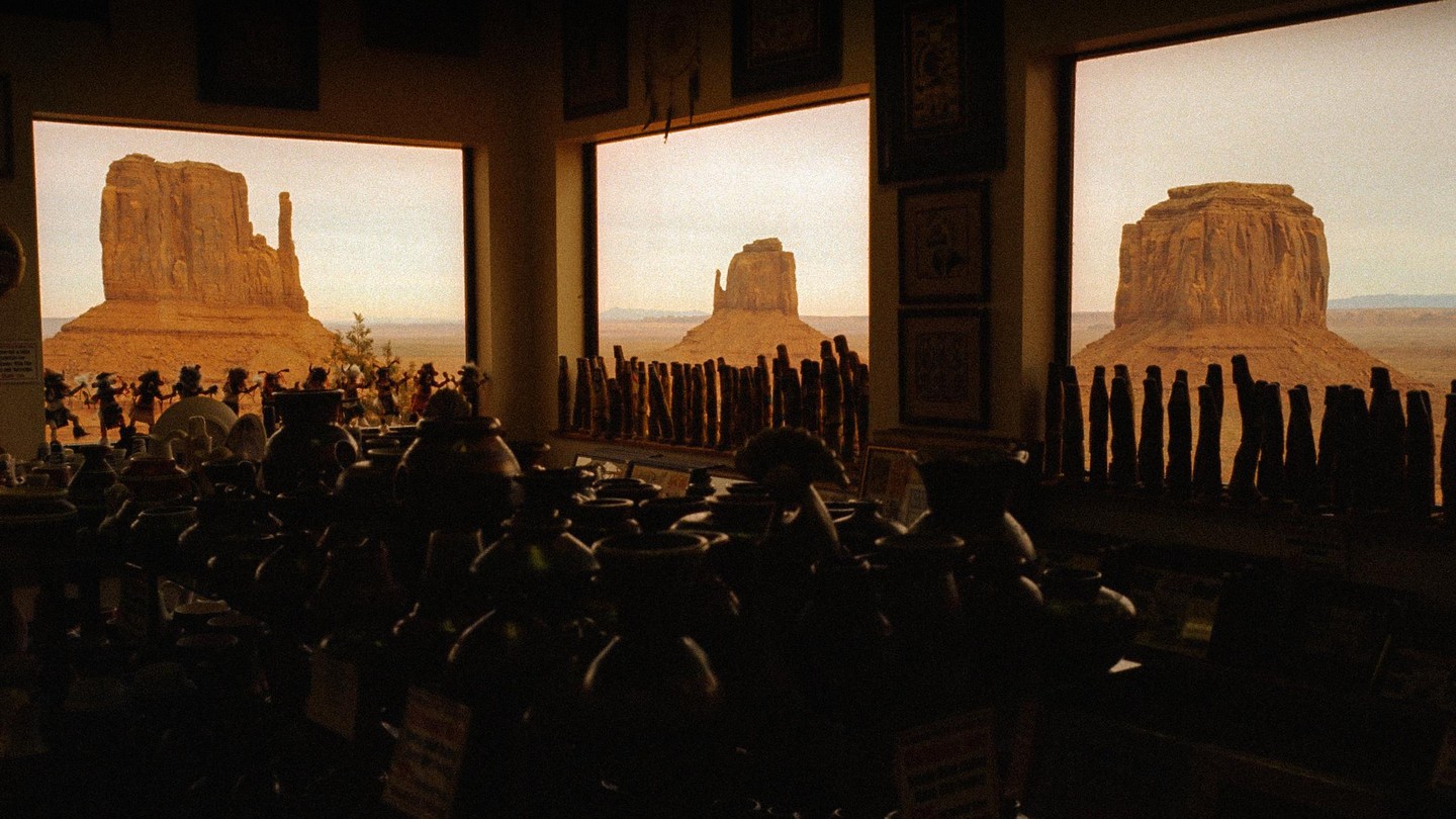Three large windows in a dark room look out onto a desert landscape with mesas and buttes.