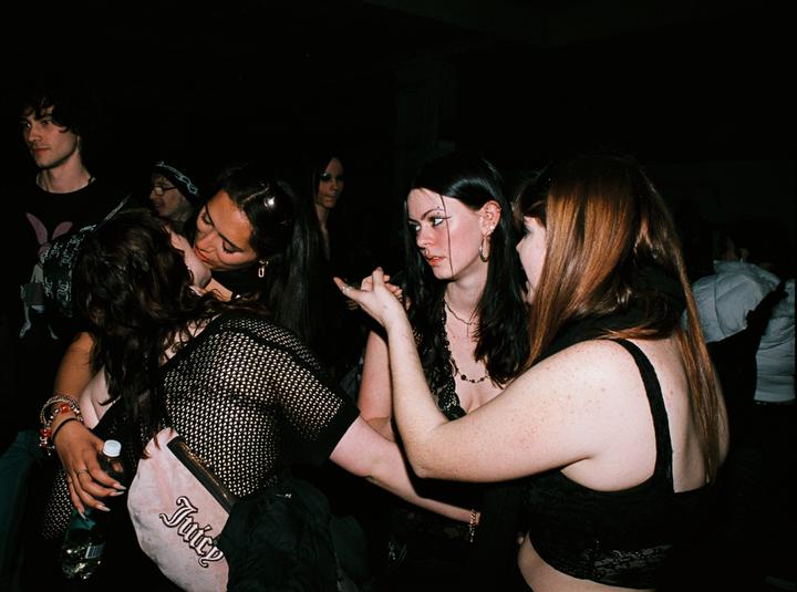 Three women are gathered closely together in a dark room, with one woman kissing another on the cheek.