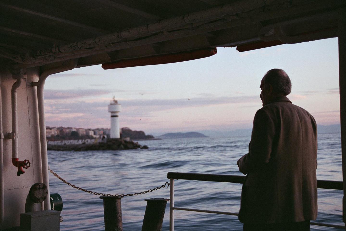 A man stands on a ferry looking out at a lighthouse and the sea at dusk.