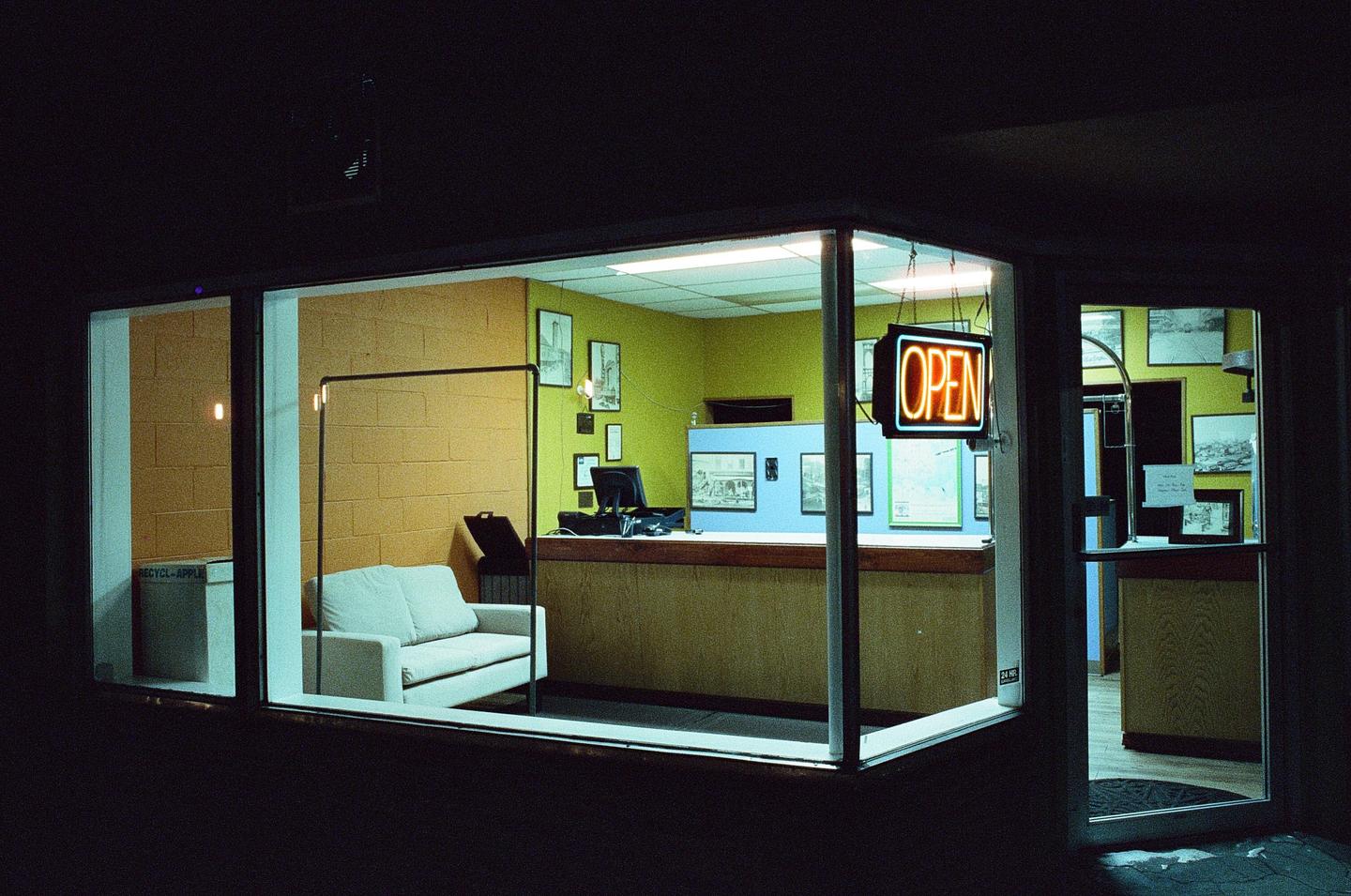 A neon "OPEN" sign hangs in the window of a business at night, revealing an interior with a couch, counter, and framed pictures.