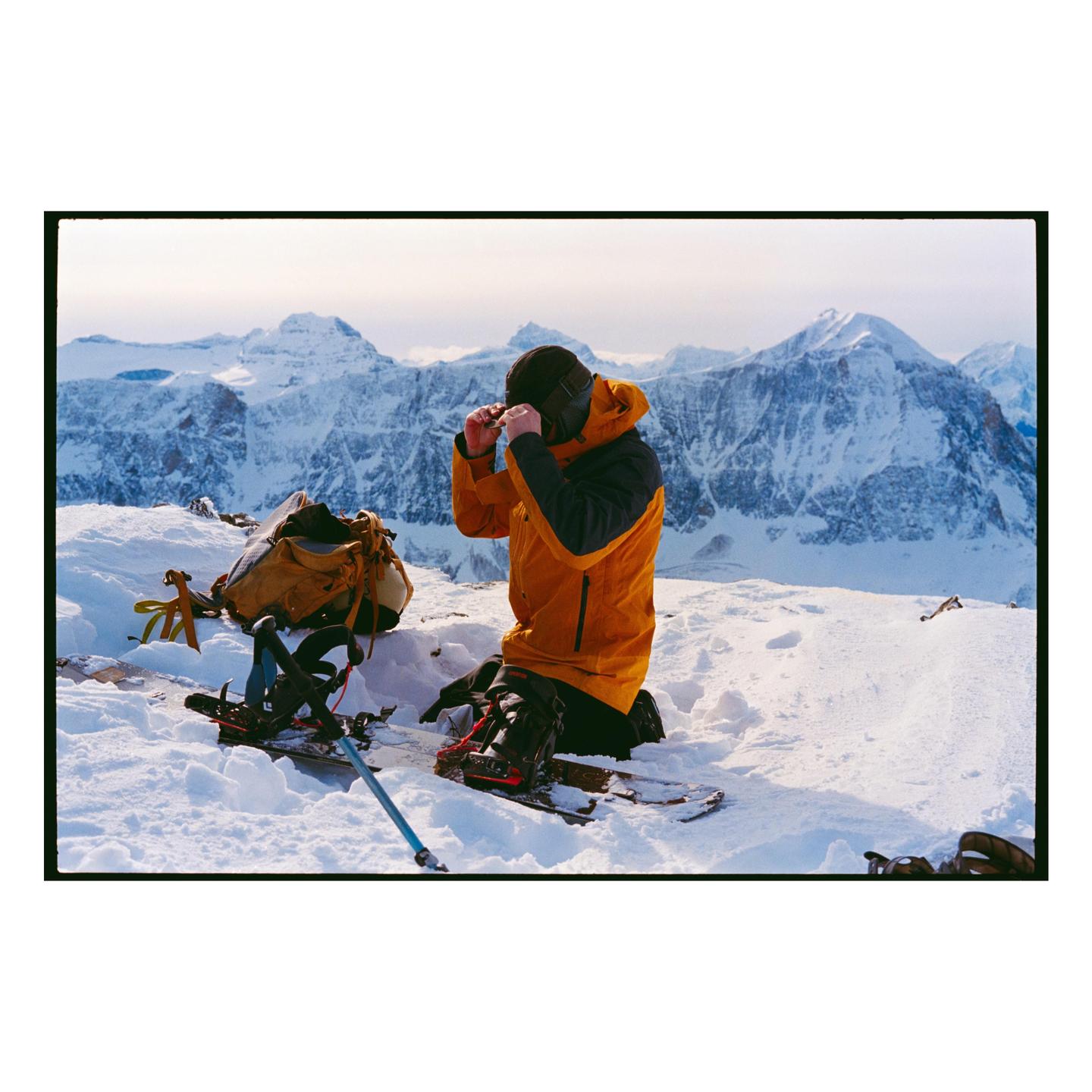 A man kneels in the snow, adjusting his goggles, with a backpack and skis beside him and snow-covered mountains in the background.