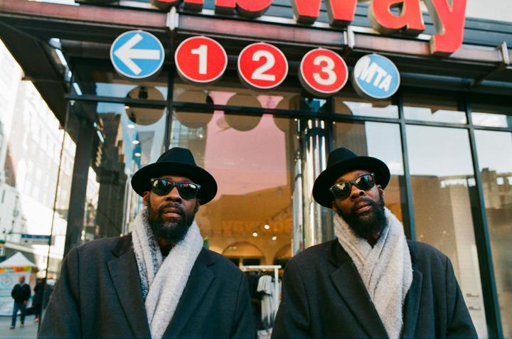 Two men wearing hats and sunglasses stand in front of a building with subway signs.