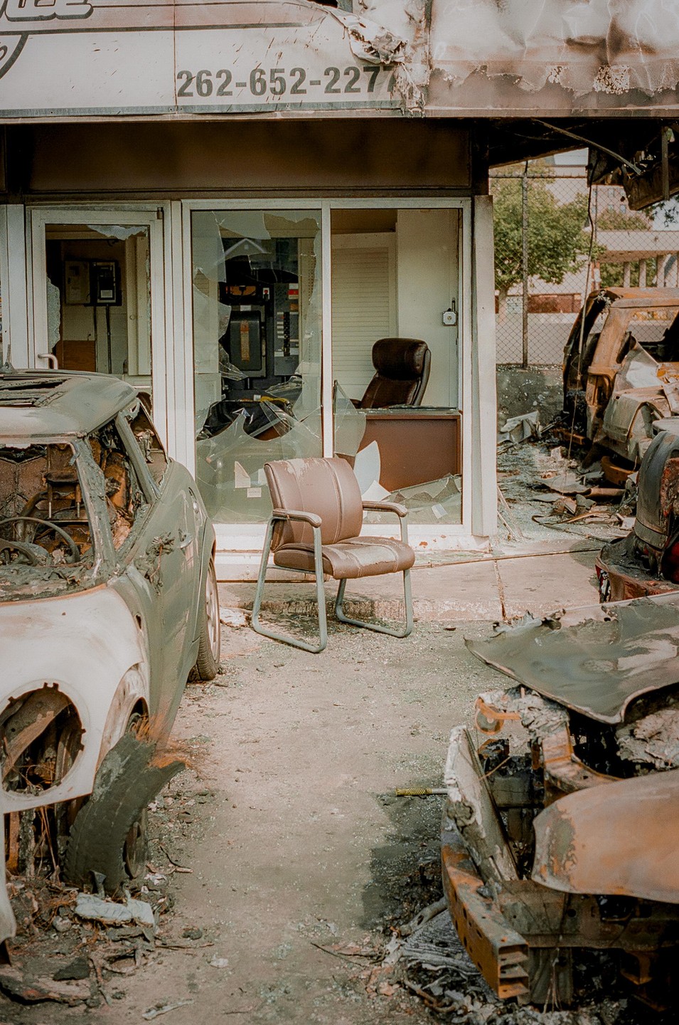 A damaged building with broken windows stands between two burnt-out cars, with a single chair in front of the entrance.