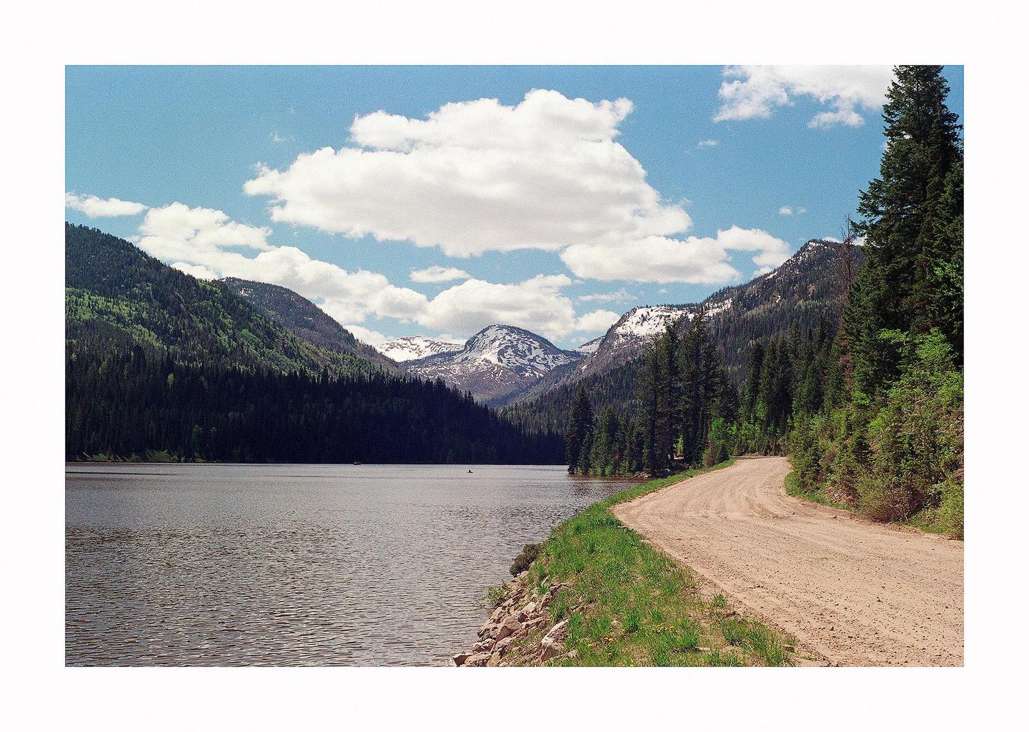 A dirt road curves along a lake with mountains and pine trees under a cloudy sky.