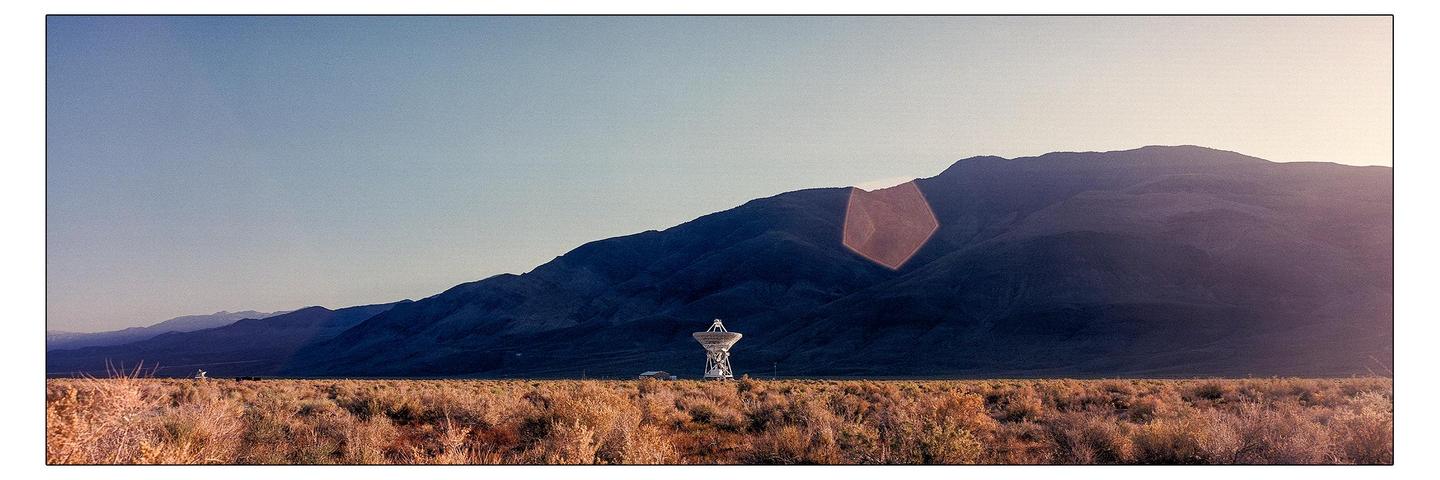 A large satellite dish stands in a field of dry brush with mountains in the background under a clear sky.
