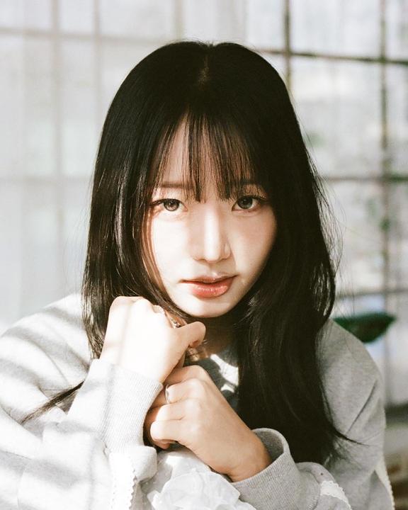 A woman with long dark hair and bangs looks directly at the viewer, with her hands clasped in front of her.