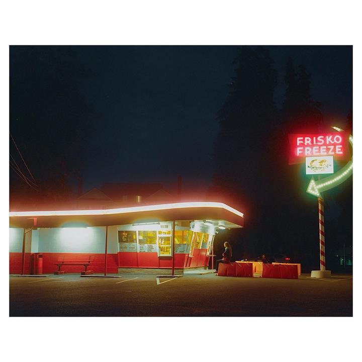 A woman sits outside a brightly lit Frisko Freeze restaurant at night.