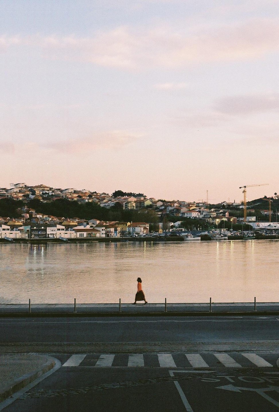 A woman walks along a waterfront promenade with a town and river in the background.