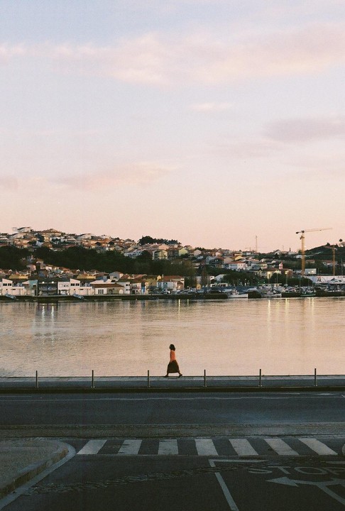 A woman walks along a waterfront promenade with a town and river in the background.