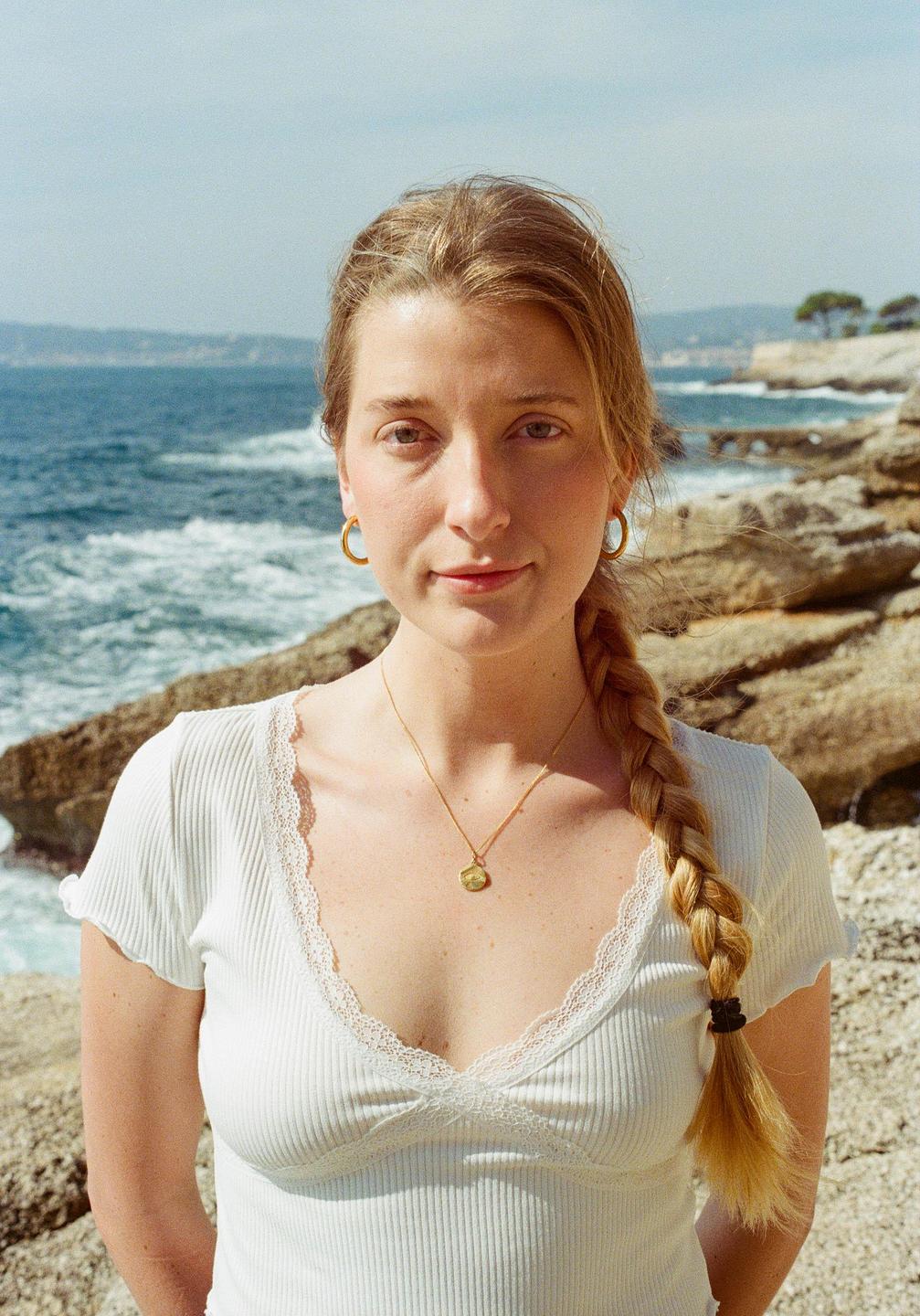 A woman with a braid and a white top stands on a rocky shore with the ocean in the background.