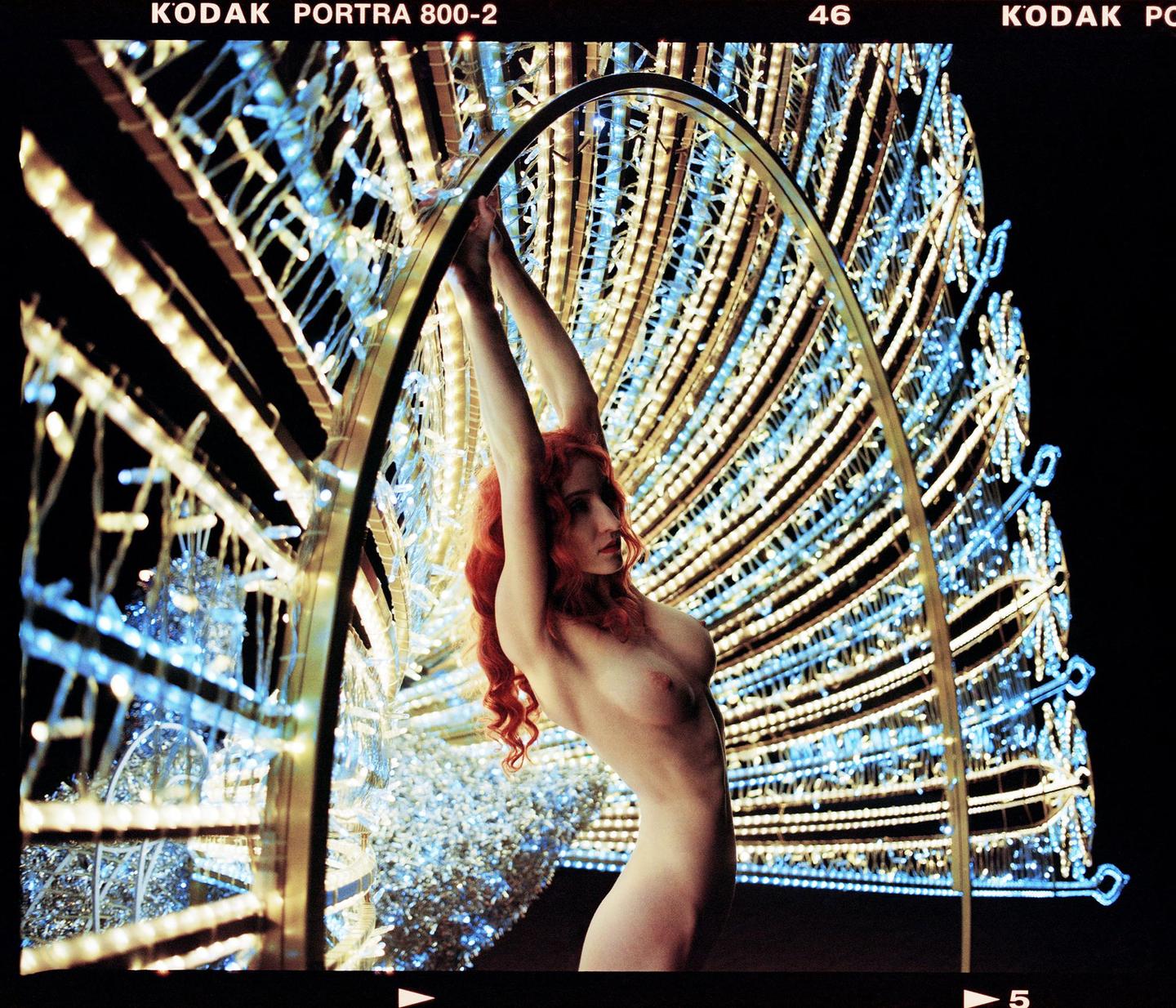 A nude woman with long red hair poses with her arms raised, framed by a large, illuminated peacock sculpture at night.