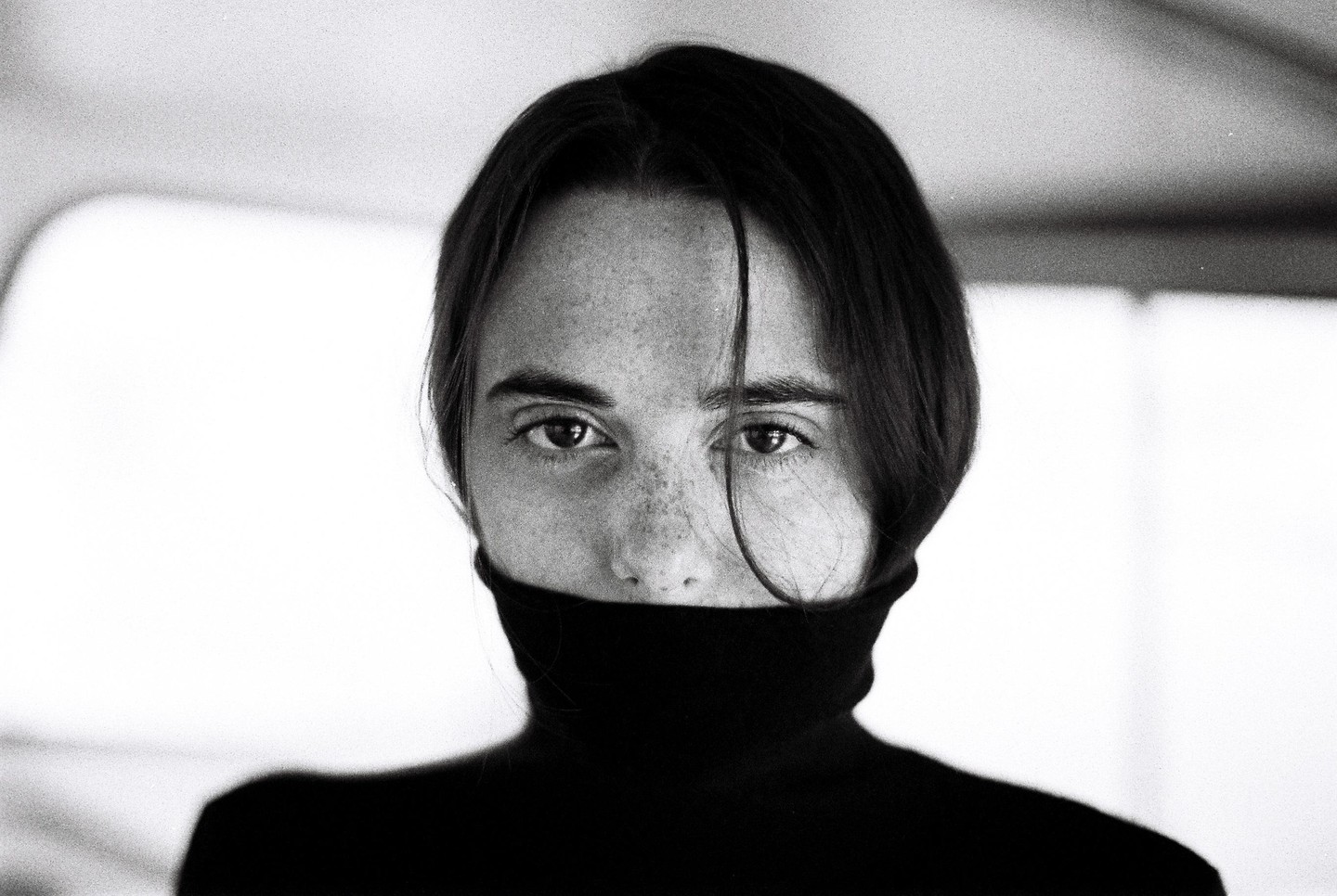 A woman with freckles and dark hair looks directly at the viewer, her face partially covered by a black turtleneck.
