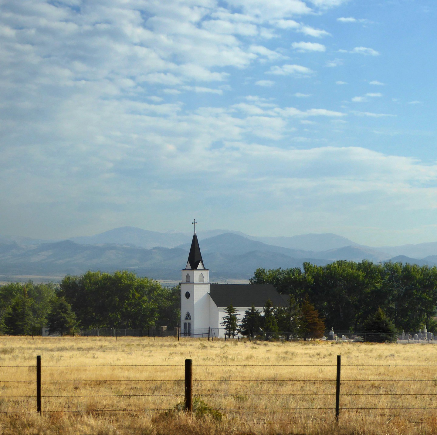 A white church with a black steeple stands in a field of dry grass with mountains in the background.