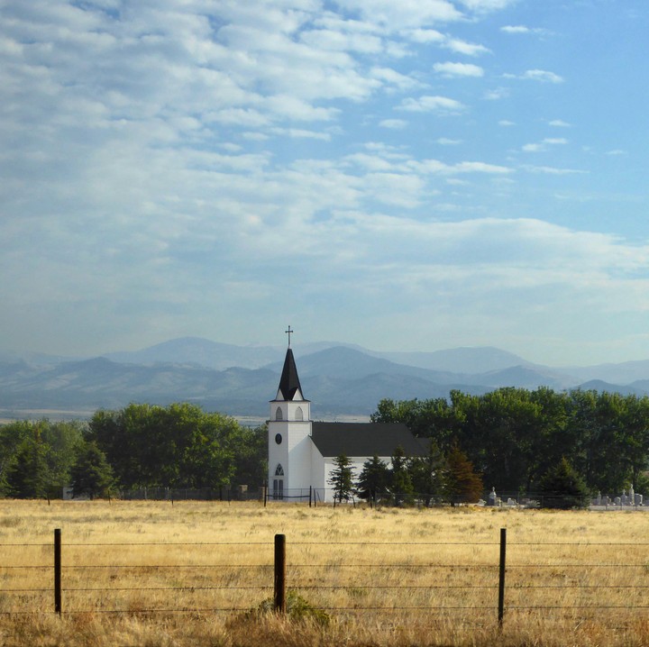 A white church with a black steeple stands in a field of dry grass with mountains in the background.