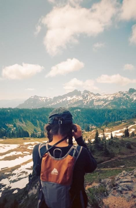 A person with a backpack and a baseball cap looks out at a mountain range with snow and a forest.
