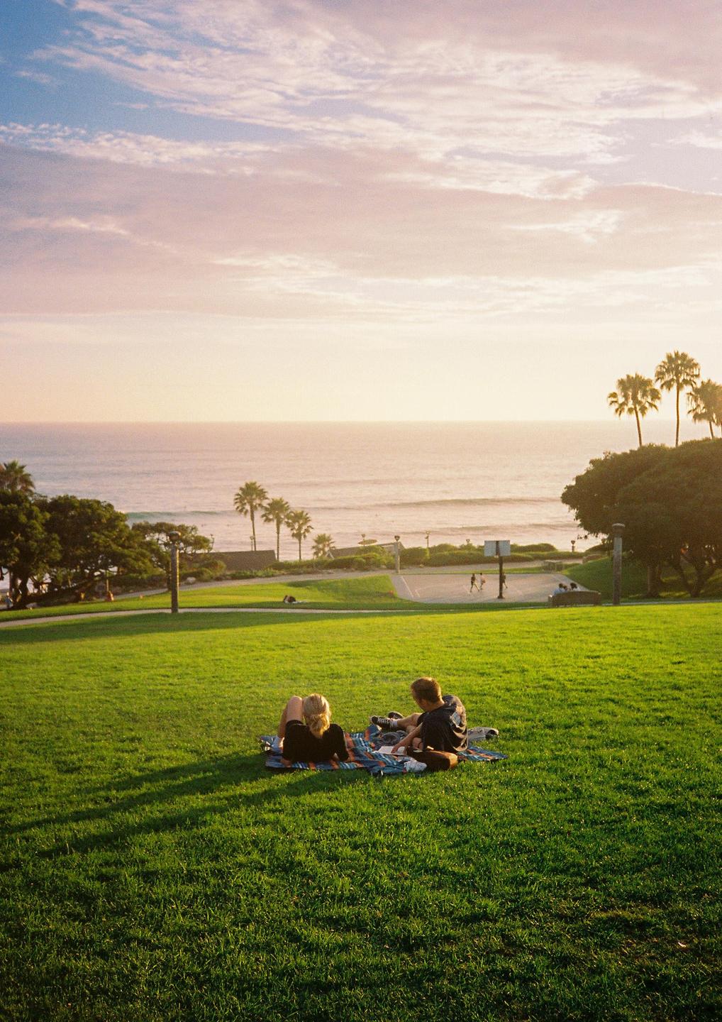 A couple sits on a blanket in a grassy park overlooking the ocean at sunset.