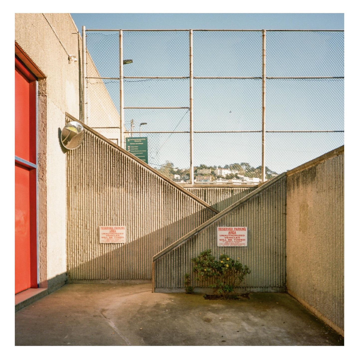 A red door and a convex mirror are next to a concrete staircase with a chain-link fence at the top.