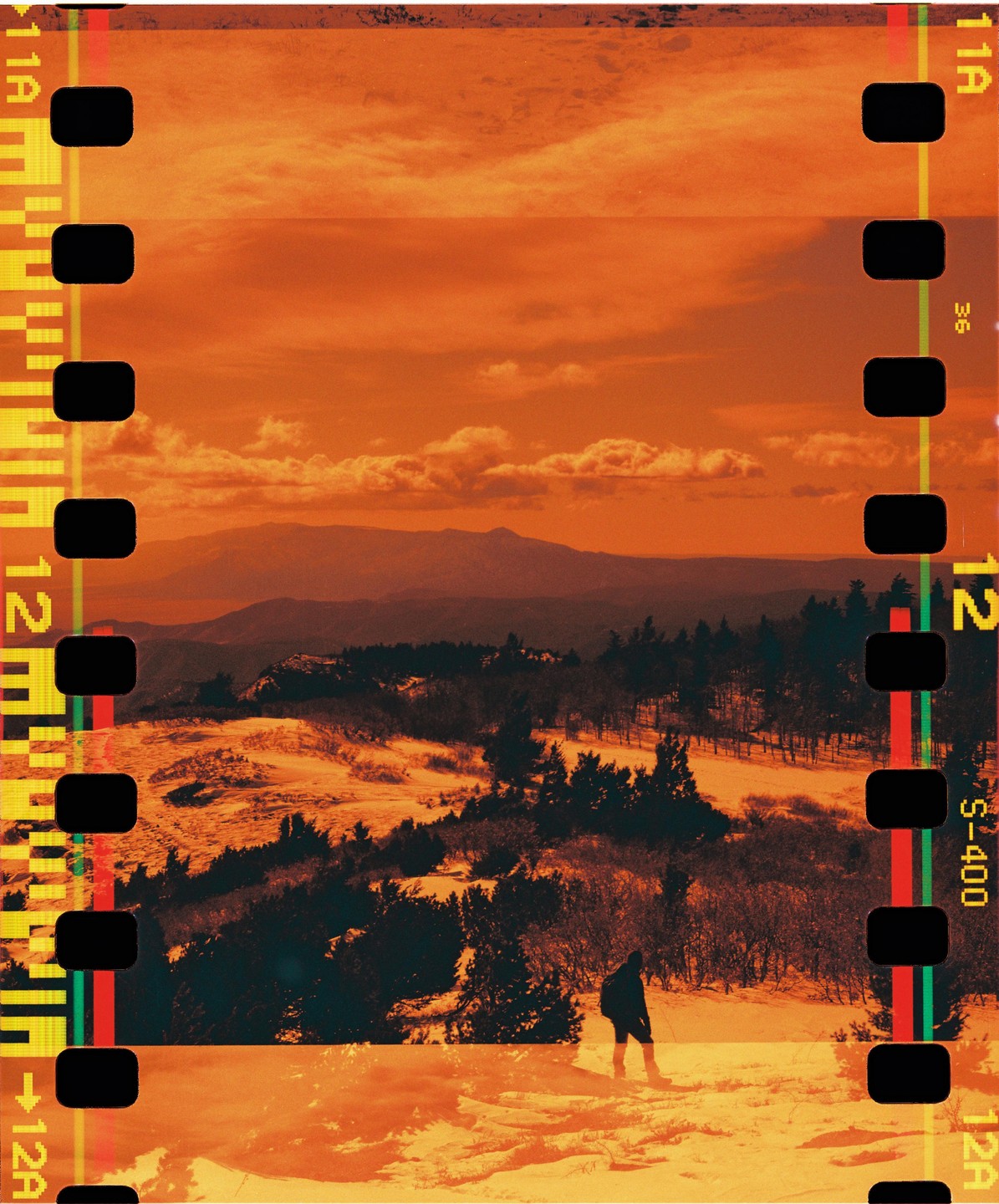A person stands on a snowy hill overlooking a forest and mountains under an orange sky.