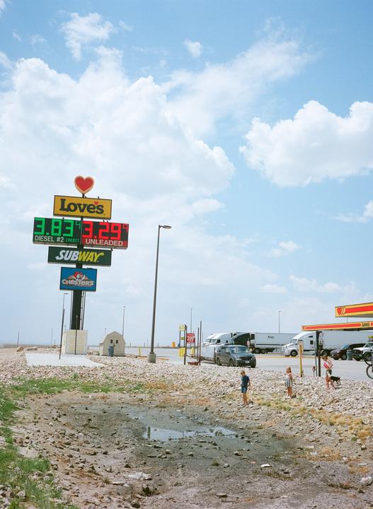 A gas station sign with prices for diesel and unleaded fuel stands next to a Love's gas station with several trucks and cars parked.