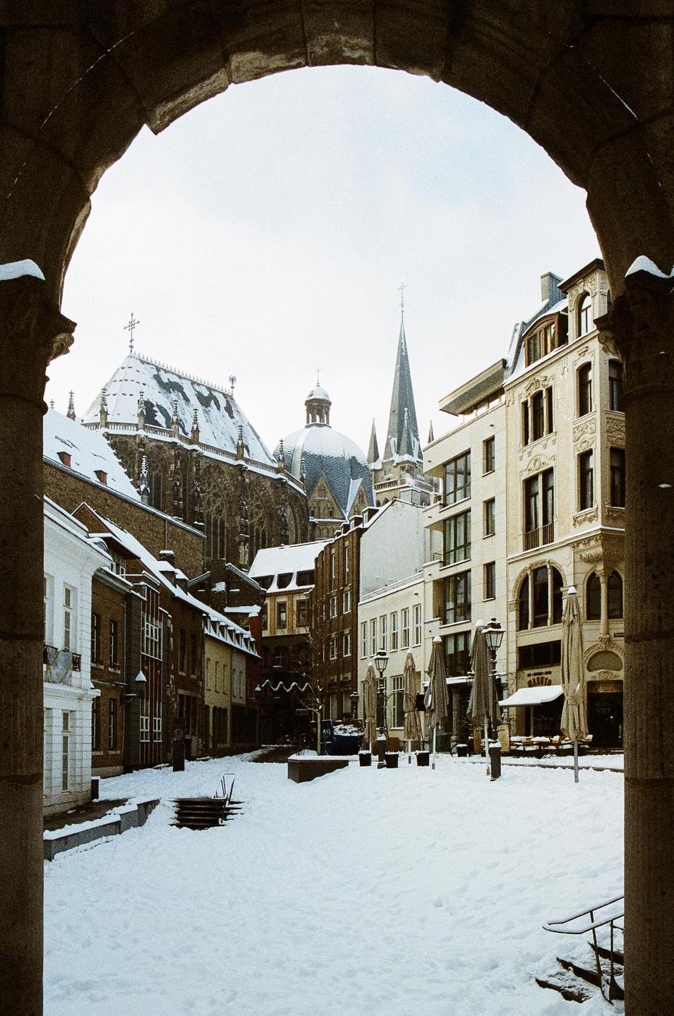 A snow-covered town square is framed by a stone archway, with historic buildings and a large cathedral visible in the background.