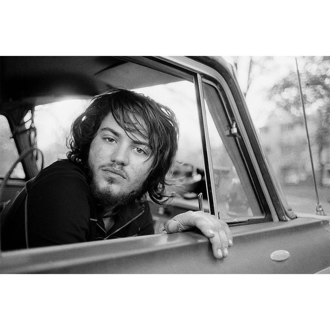 A man with long hair and a beard looks out the window of a car.