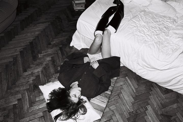 A woman lies on a pillow on a wooden floor next to a bed, wearing a coat and boots.