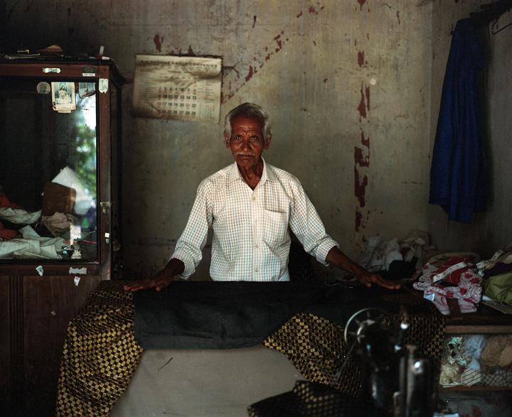 An elderly man with a mustache and white hair stands behind a table with fabric in a room with peeling paint.