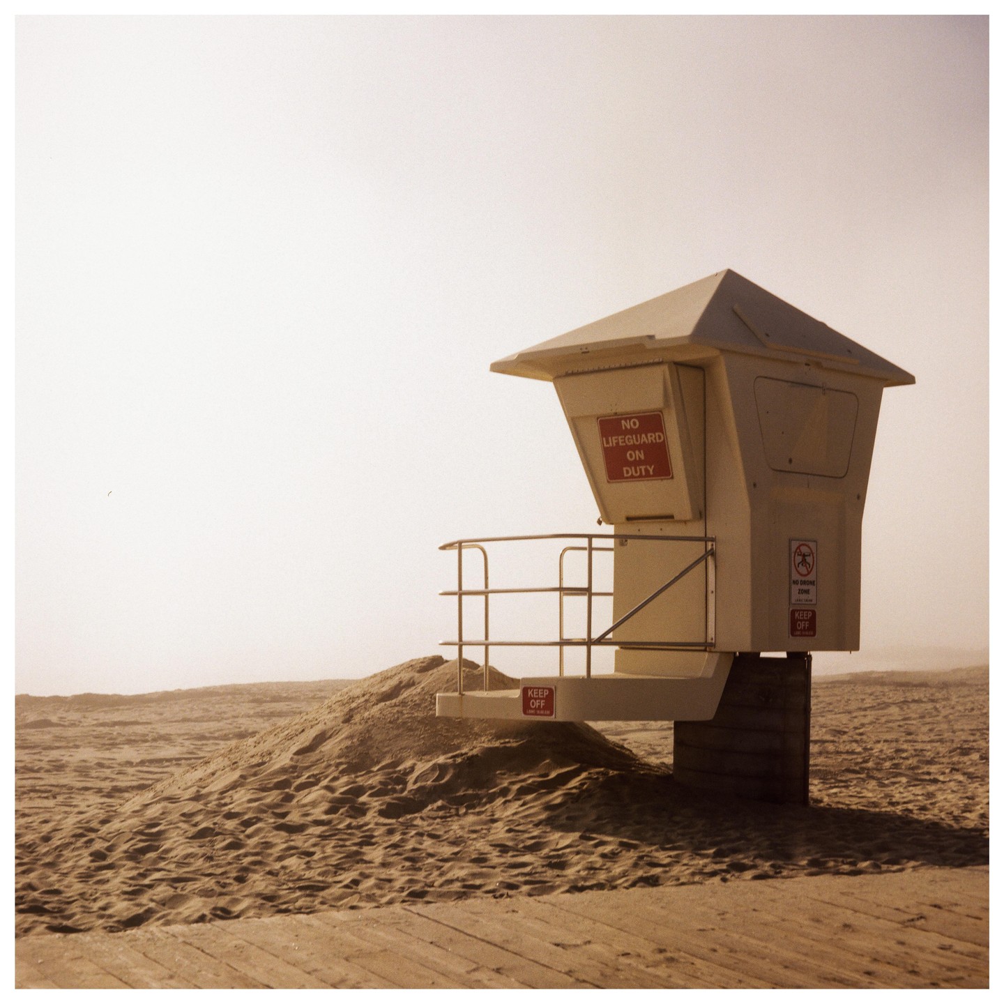 A lifeguard tower stands on a sandy beach with a wooden boardwalk in the foreground under a hazy sky.