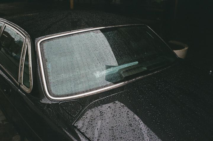 Raindrops cover the windshield and hood of a dark car.