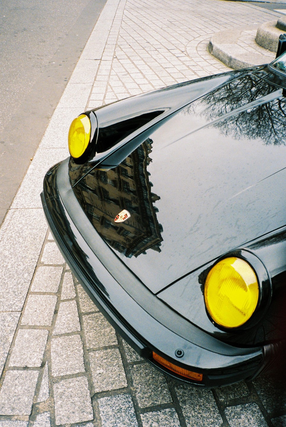 A black car with yellow headlights is parked on a cobblestone street, reflecting a building and trees.