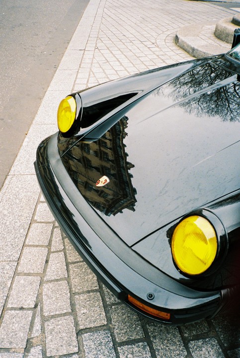 A black car with yellow headlights is parked on a cobblestone street, reflecting a building and trees.
