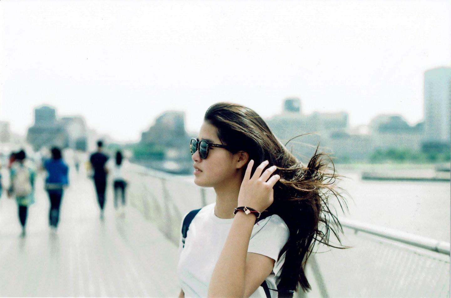 A young woman with sunglasses and long hair blowing in the wind stands on a bridge with people and buildings in the background.