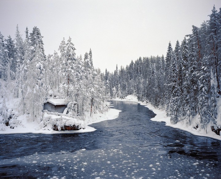 A log cabin sits on the snowy bank of a winding river lined with snow-covered pine trees.