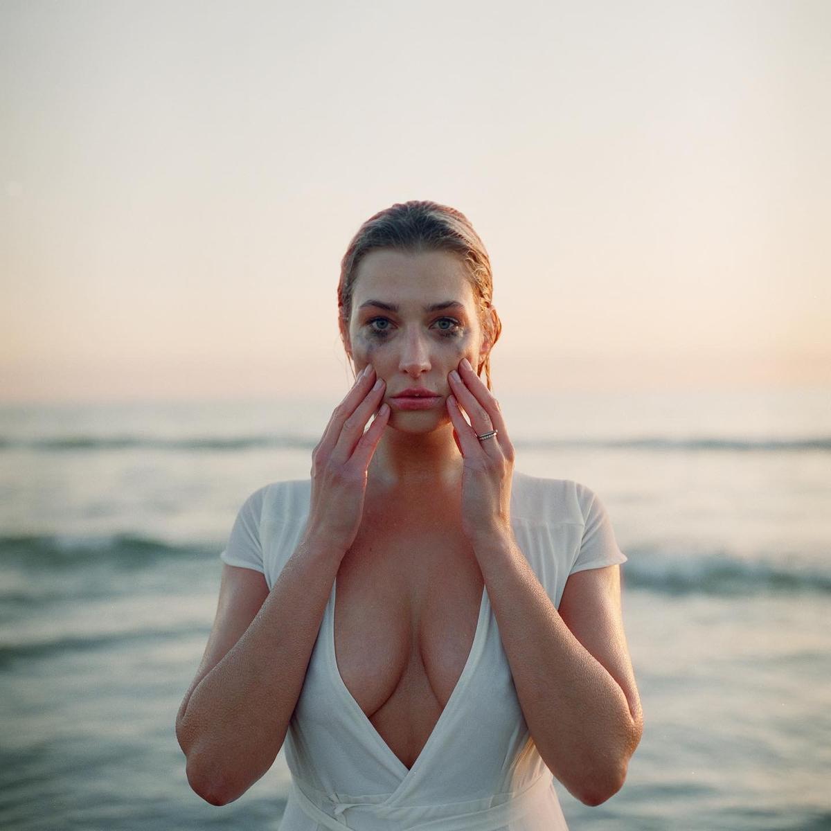 A woman with smudged makeup on her face stands in front of the ocean with her hands on her cheeks.