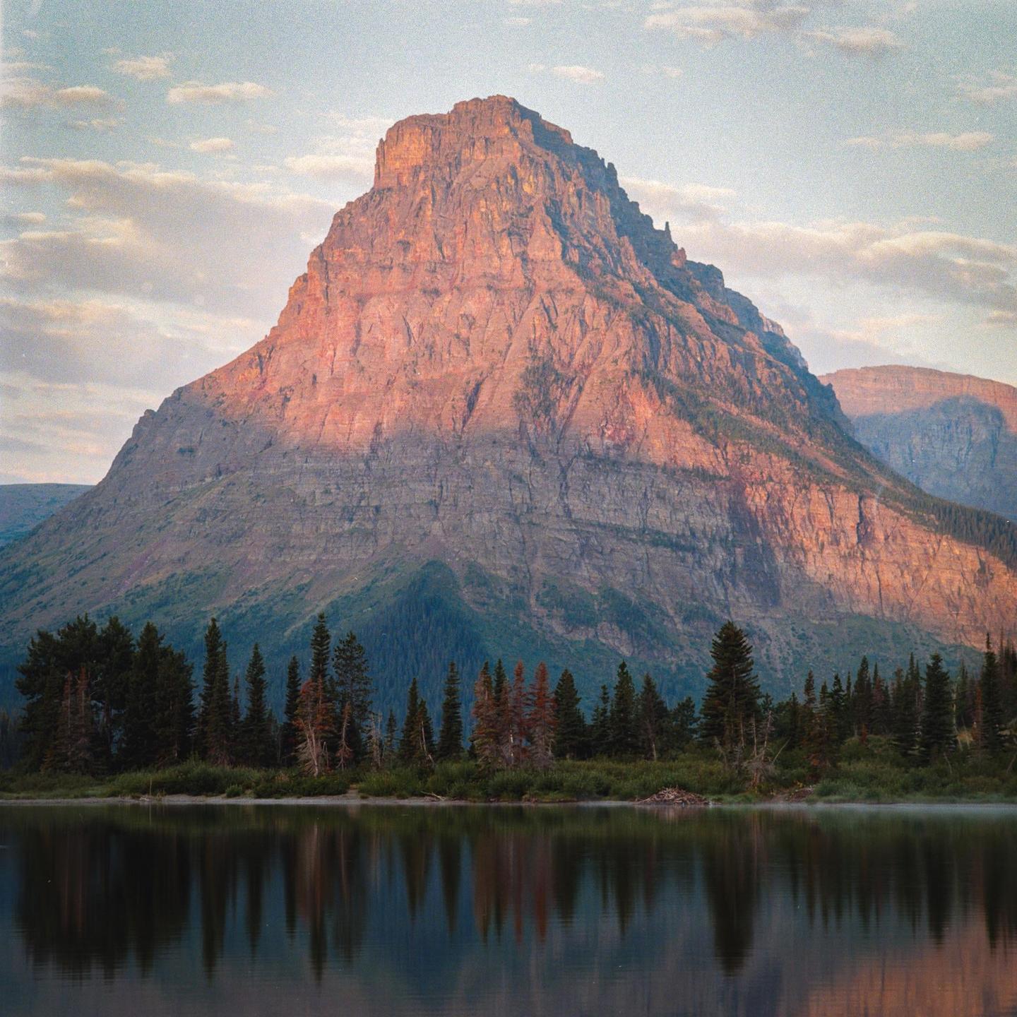A large mountain with a forested base is reflected in a lake under a cloudy sky.