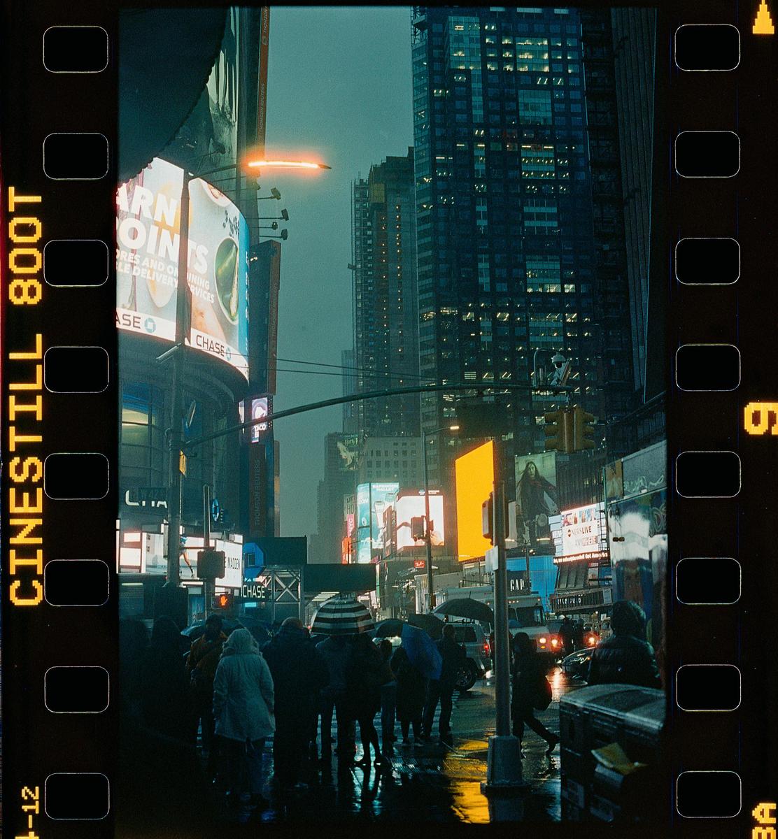 A crowd of people with umbrellas walk on a wet street at night, surrounded by tall buildings and bright billboards.