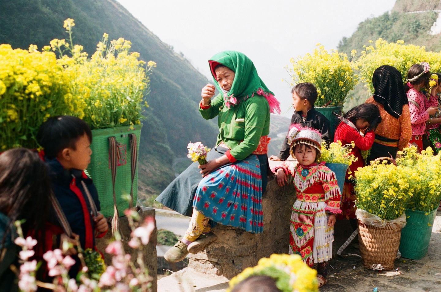 A woman and several children are gathered outdoors among baskets of yellow flowers with a mountain in the background.